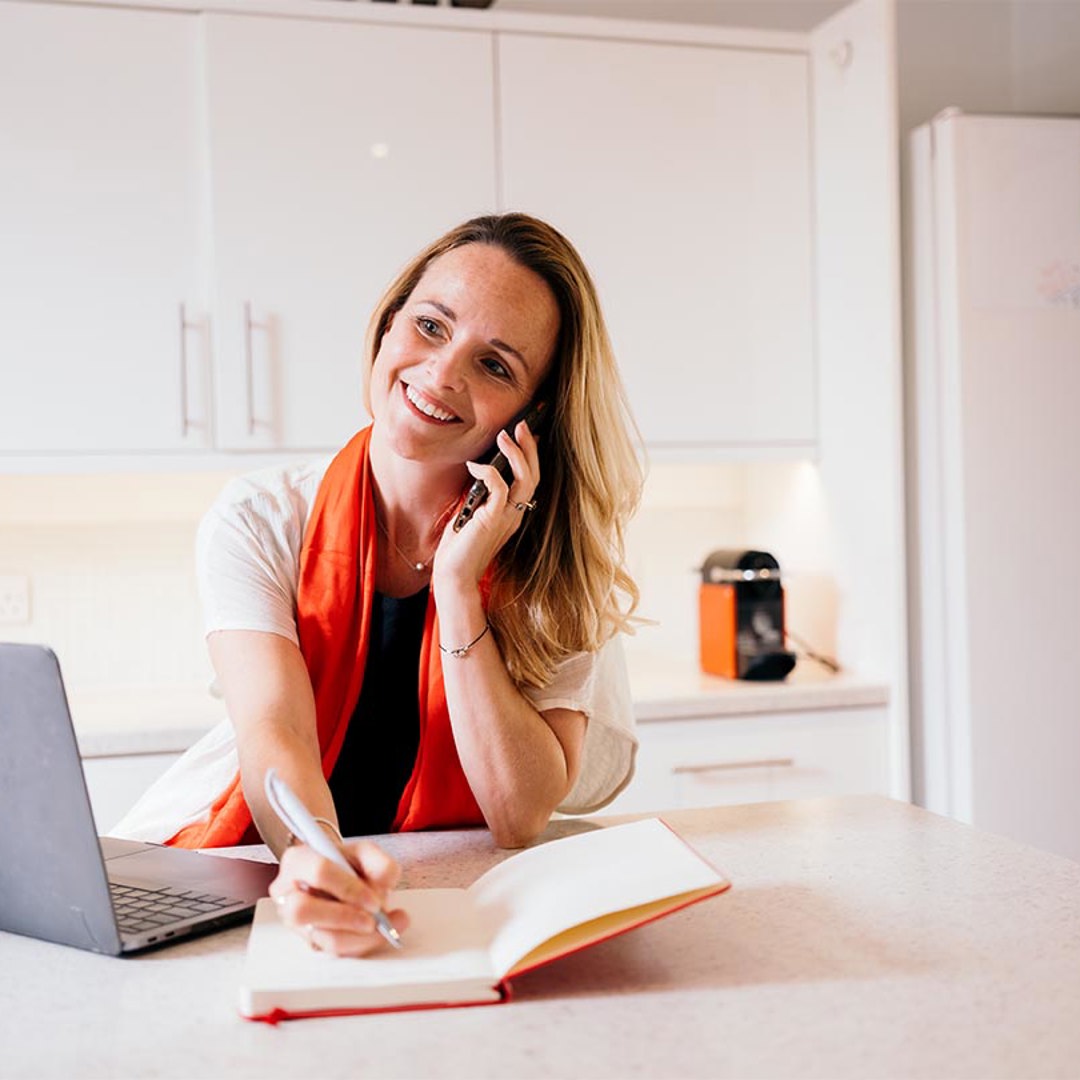 Woman on mobile phone in front of lap top writing in note book
