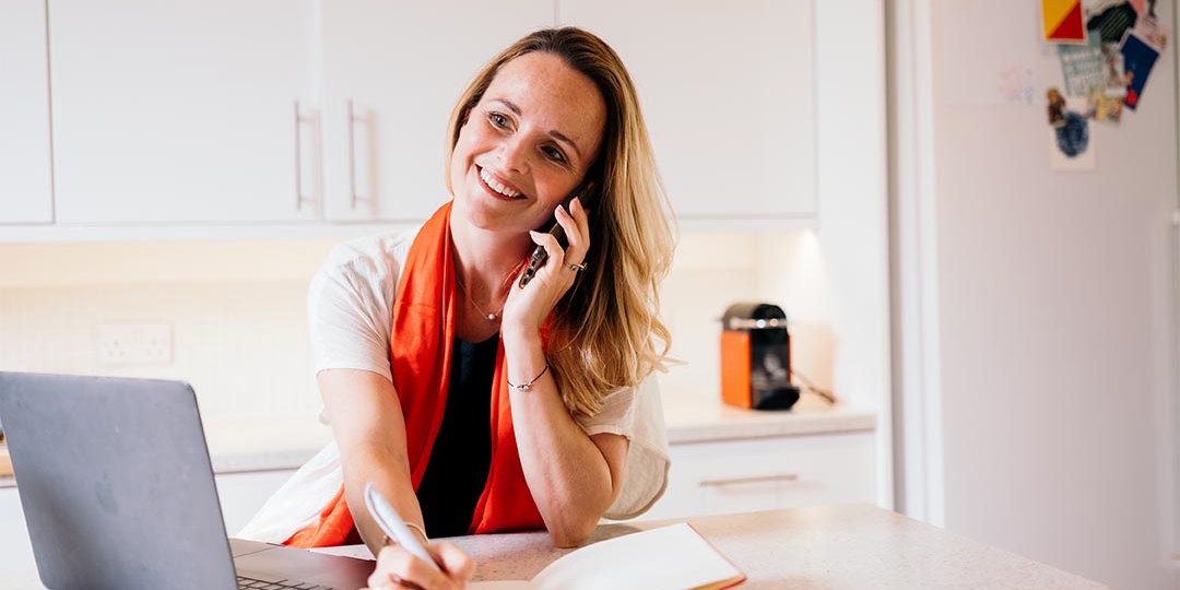 Woman on mobile phone in front of lap top writing in note book