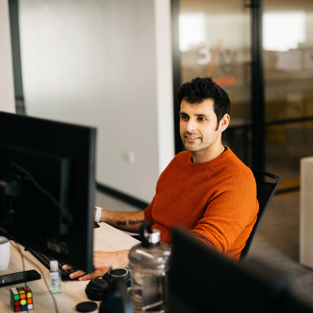 Man in orange jumper sat at desk in front of computer screens