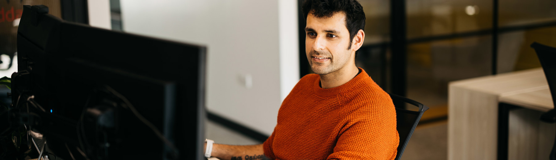Man in orange jumper sat at desk in front of computer screens